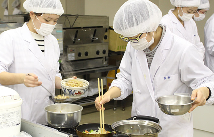 厨房で白衣を着た人たちが料理の盛り付けや調理作業をしている様子の写真