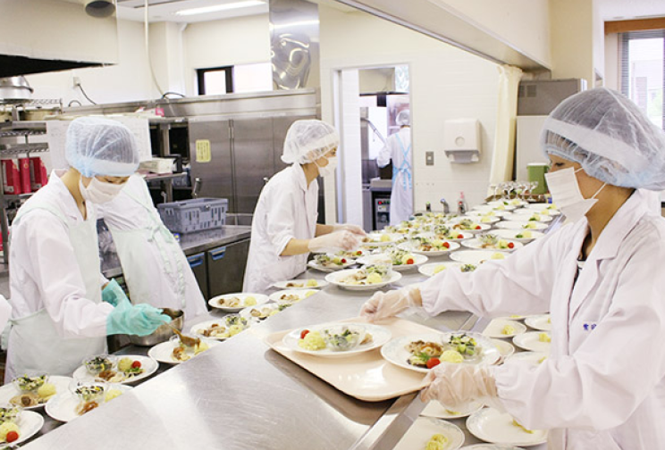 厨房で白衣を着た人たちが多数の料理を並べて準備している様子の写真