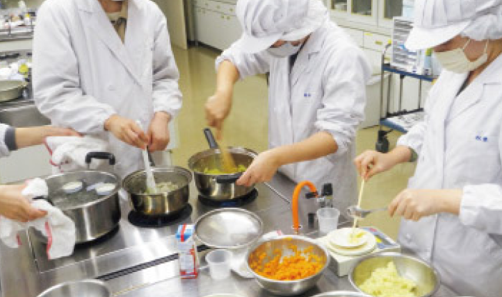 厨房で白衣を着た人たちが複数の鍋を使って調理作業をしている様子の写真