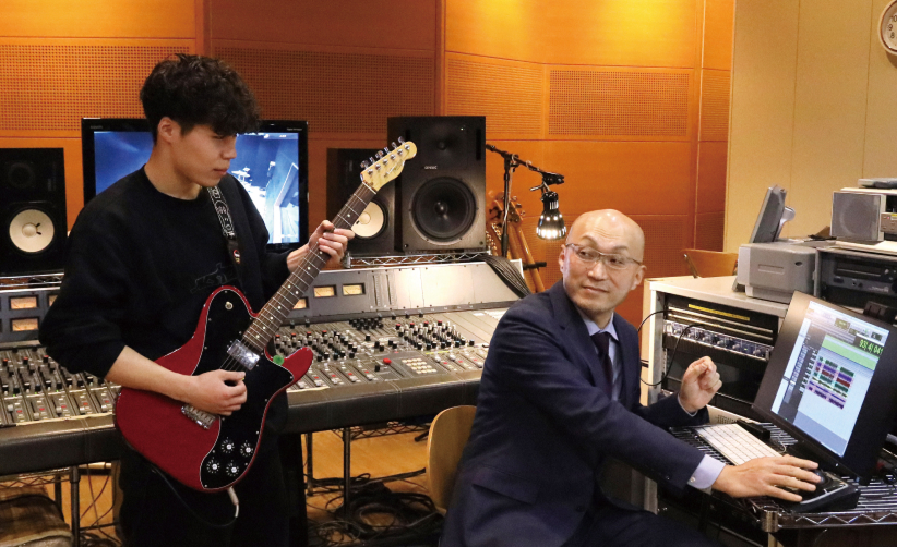 音響スタジオでギター演奏と音響機器の操作が同時に行われている様子の写真